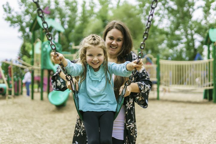 Mom in park pushing her young daughter on a swing.