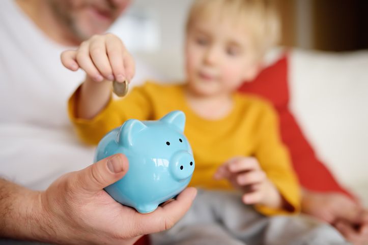 Father and young son saving money by putting a coin in a blue piggy bank.